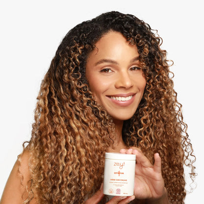 Femme souriante aux cheveux bouclés, tenant un pot de crème sans rinçage "Definition 1" de Zeyt. Le produit nourrit, définit et fixe les boucles grâce à des ingrédients comme l'aloe vera et le beurre de mangue.