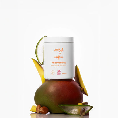 Image de la Crème sans Rinçage Definition de la marque Zeyt. Le pot est posé sur un fond blanc avec des morceaux de mangue, d'aloe vera et d'autres ingrédients naturels. Ce produit nourrit, définit et fixe les boucles, et est certifié biologique par Cosmos Organic.