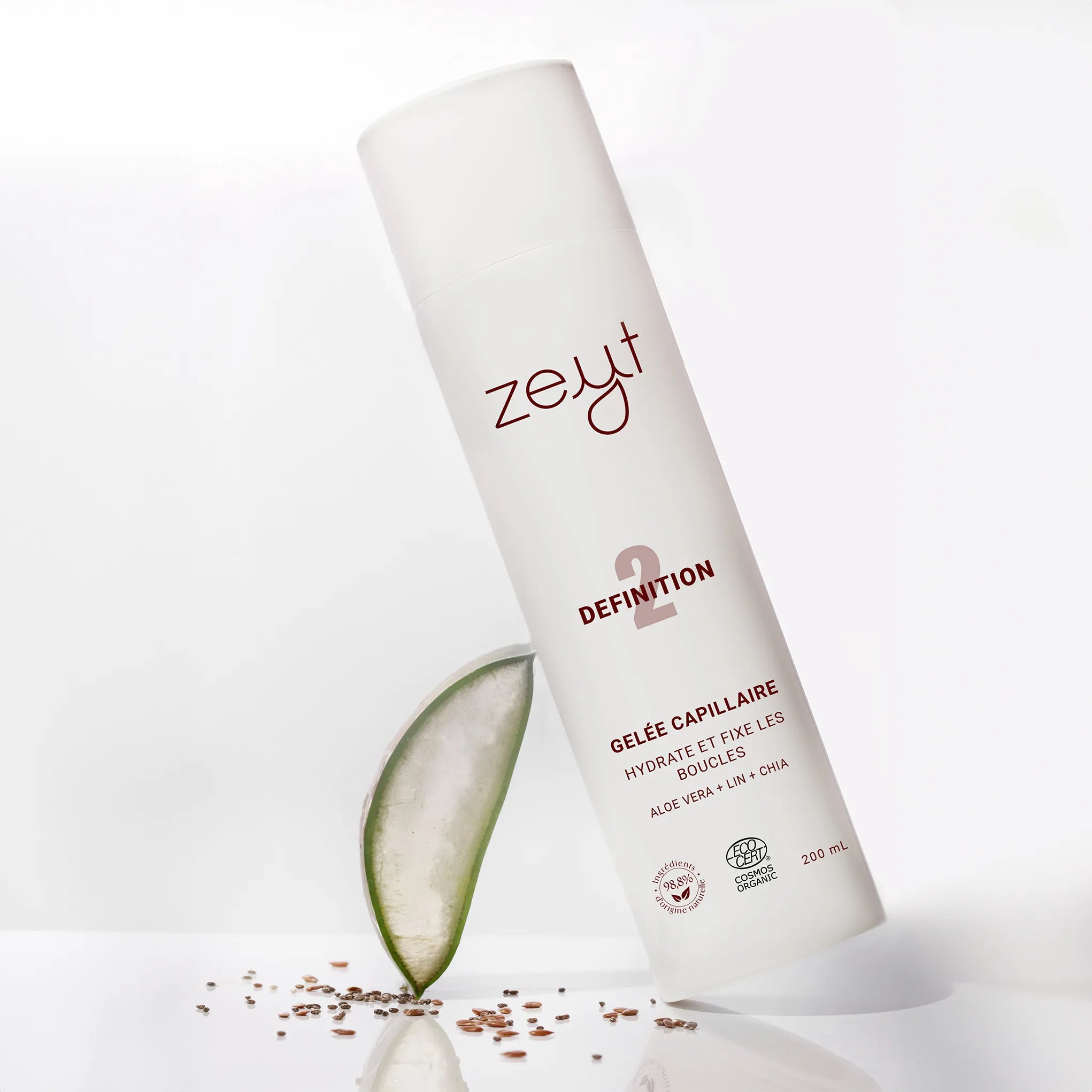 Image de la Gelée Capillaire Definition de la marque Zeyt. Le flacon est incliné sur un fond blanc avec un morceau d'aloe vera et des graines de lin. Ce produit hydrate et fixe les boucles, avec des ingrédients tels que l'aloe vera, le lin et le chia, et est certifié biologique par Cosmos Organic.