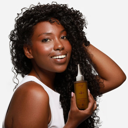 Femme souriante aux cheveux bouclés, tenant une bouteille de sérum cheveux secs "Definition 3" de Zeyt. Le sérum nourrit et scelle l'hydratation des cheveux grâce à des ingrédients comme l'argan, le moringa et le ricin.