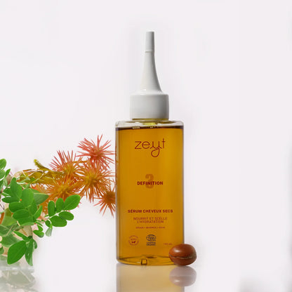 Image du Sérum Cheveux Secs Definition de la marque Zeyt. Le flacon est debout sur un fond blanc avec une plante et une noix de ricin à ses côtés. Ce sérum nourrit et scelle l'hydratation des cheveux secs, avec des ingrédients comme l'argan, le moringa et le ricin, et est certifié biologique par Cosmos Organic.